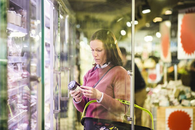 Mature woman holding bottle by refrigerator in supermarket