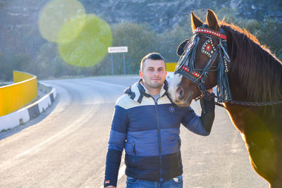 Portrait of smiling mid adult man touching horse while standing on road during sunset