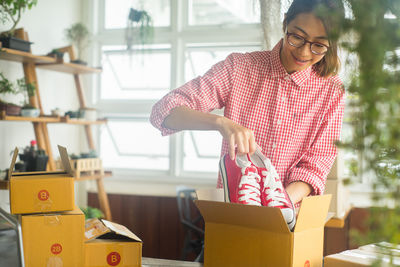 Smiling young woman removing shoes from cardboard box on table at home