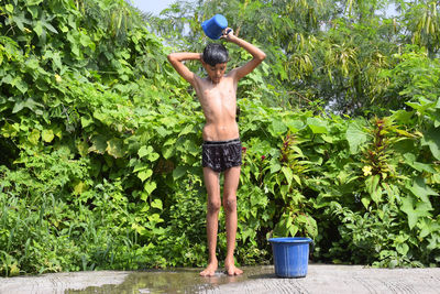 A rural child taking a bath by the bucket on the road, plants and greenery in the background