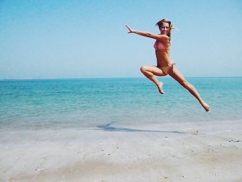 Full length of woman in bikini jumping over shore at beach against clear blue sky