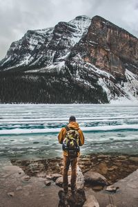 Rear view of man with backpack standing on rock by lake against mountain during winter