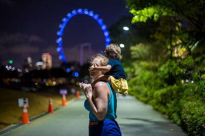 Mother carrying daughter against illuminated ferris wheel