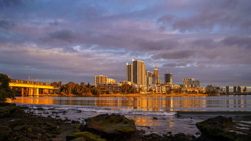 Illuminated buildings by river against sky during sunset