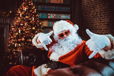 Portrait of man in santa claus costume gesturing while sitting by christmas tree at home