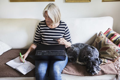 Senior woman writing in book while using laptop and credit card by dog on sofa at home