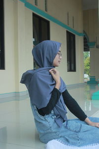 Young woman wearing hijab sitting against wall