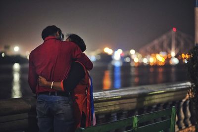 Rear view of man standing by railing against illuminated city at night