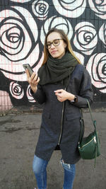 A young woman in glasses, a gray coat, a green scarf and jeans looks at the smartphone screen