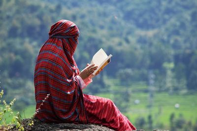 Teenage girl wearing hijab reading book while sitting on rock