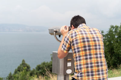 Man using stationary binoculars  on a hill against the background of the mountains and sea