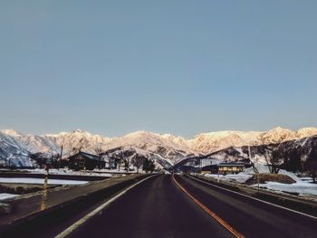 Empty road leading towards snowcapped mountains against clear blue sky during sunset