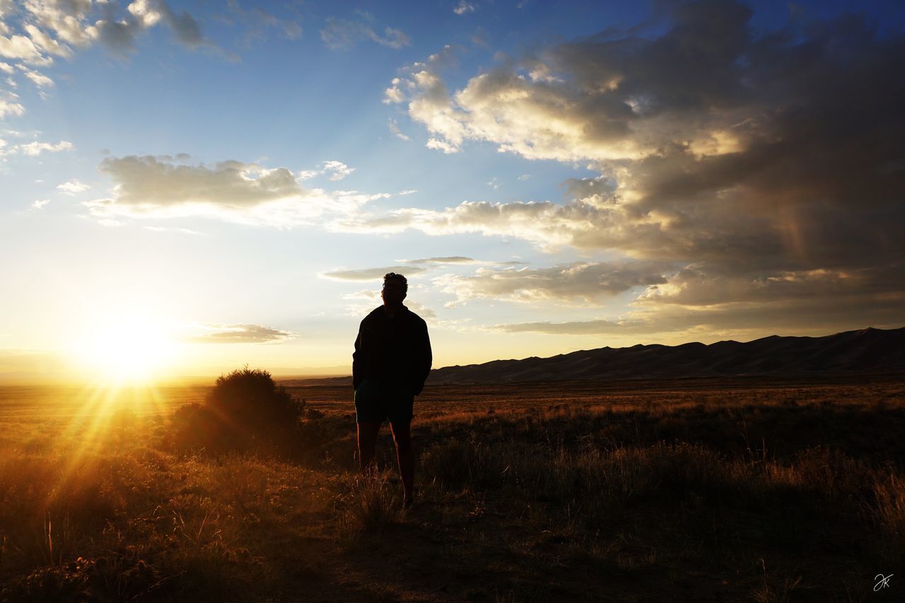 REAR VIEW OF SILHOUETTE MAN STANDING ON FIELD DURING SUNSET