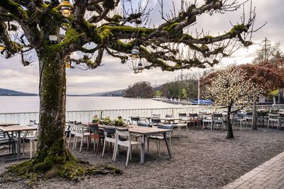 Empty chairs and table by river against sky