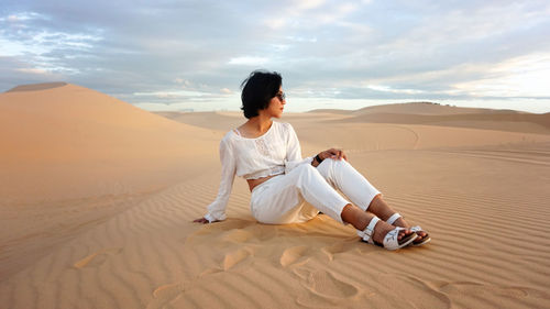 Full length of woman sitting on sand dune against sky