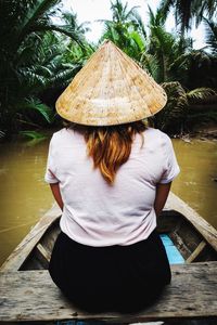 Rear view of woman with hat sitting by tree