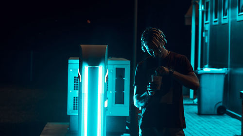 Man having drink while standing by illuminated lighting equipment