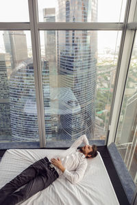 Businessman with a beard and clothes is lying on bed in a skyscraper apartment during the summer day