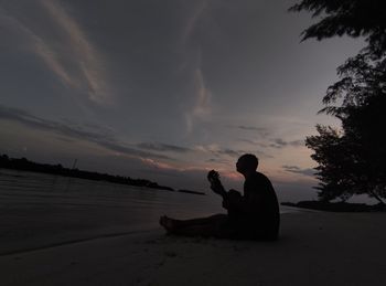 Silhouette man sitting on beach against sky during sunset