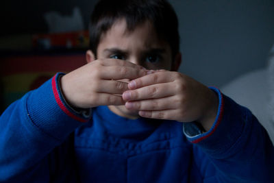 Portrait of boy with hands covering face sitting against wall at home