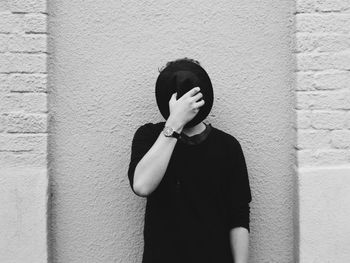 Teenage boy covering face with hat while standing against wall