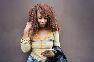 Young woman using phone while standing against wall