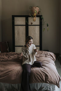 Smiling non-binary person talking through mobile phone sitting on bed at home