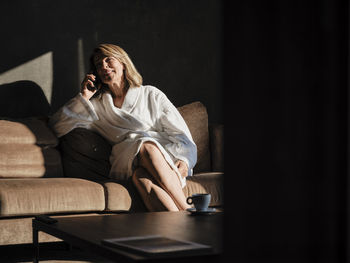 Smiling senior woman talking on phone while sitting on sofa in hotel room