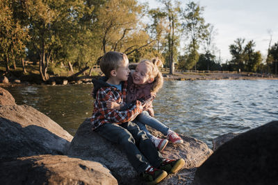 Happy siblings sitting on rock by lake simcoe during sunset