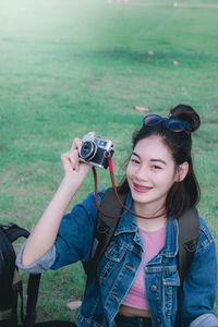 Portrait of smiling young woman holding camera while sitting on grassy field