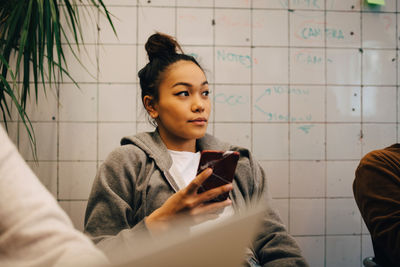 Young female programmer holding smart phone while sitting amidst colleagues against wall at office