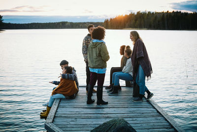 Female and male friends talking on pier over lake against sky during sunset