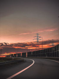 Road by silhouette electricity pylons against sky during sunset