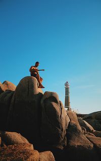 Low angle view of mid adult man playing guitar while sitting on rock formation against blue sky