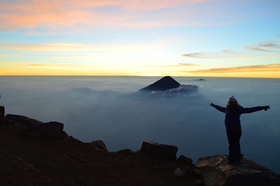 Silhouette woman with arms outstretched standing on cliff against sky during sunset