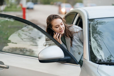 Beautiful girl with long hair in a grey trench coat using smartphone call gets into the car