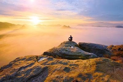 Hiker in squatting position on peak of rock and watching in colorful mist and fog in morning valley