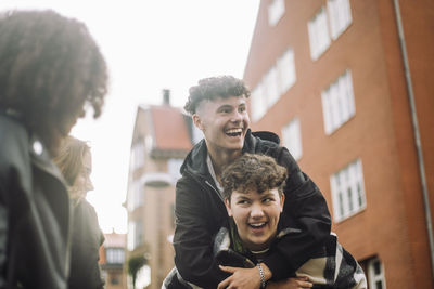 Low angle view of happy teenage boy piggybacking male friend in front of building