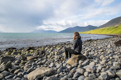 Rear view of woman sitting on pebbles at beach against sky