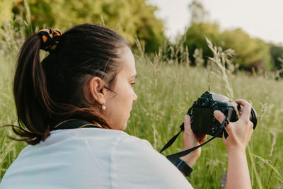 Young woman crouching in meadow, using camera and taking photos of plants and flowers