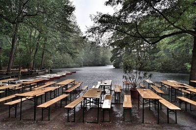 Empty chairs and tables by river in forest against sky