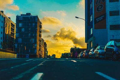 Road by buildings against sky during sunset
