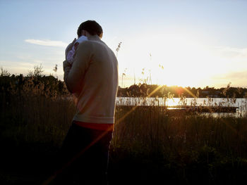 Rear view of man standing by lake against sky during sunset