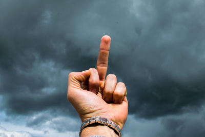 Cropped hand of person showing middle finger against cloudy sky