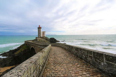 Pier leading towards lighthouse by sea against cloudy sky