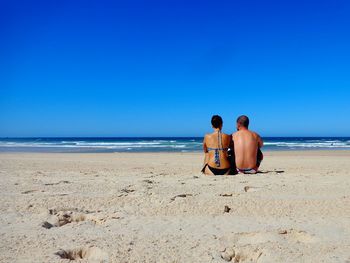 Couple sitting at beach against clear blue sky