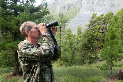 Man looking through binoculars in forest against trees
