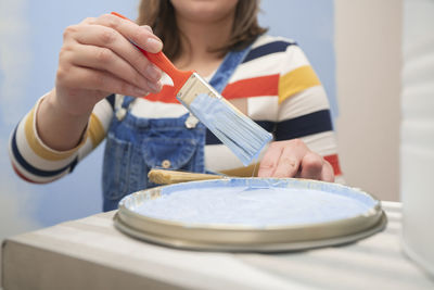 Close up of woman's hand wearing overalls, taking out a brush with blue paint from a tray with paint