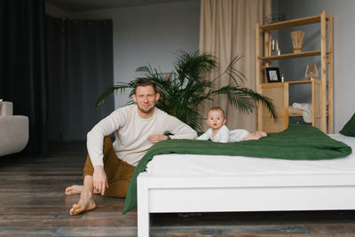 A young dad is sitting next to the bed on which his baby son is lying