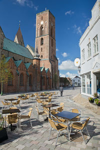 Chairs and tables at sidewalk cafe by buildings in city against sky